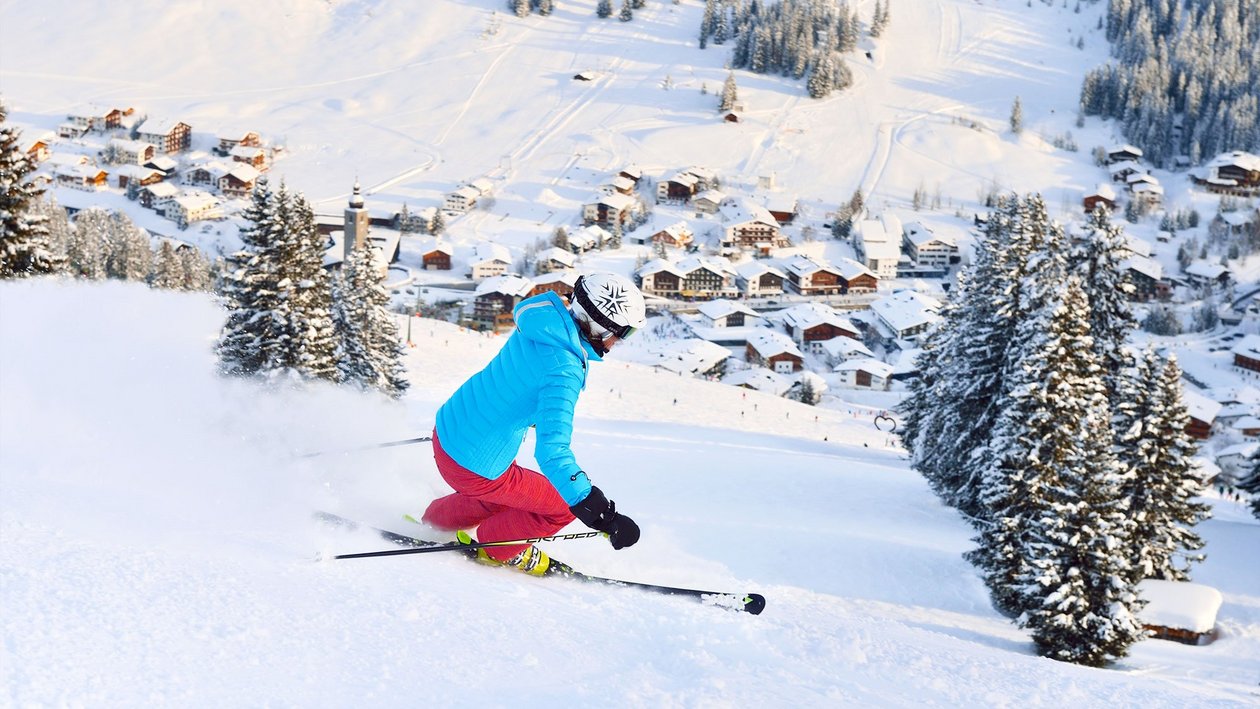 Skifahren in den Alpen Skifahrer fährt eine verschneite Abfahrt mit Bergresort im Hintergrund entlang des Hangs