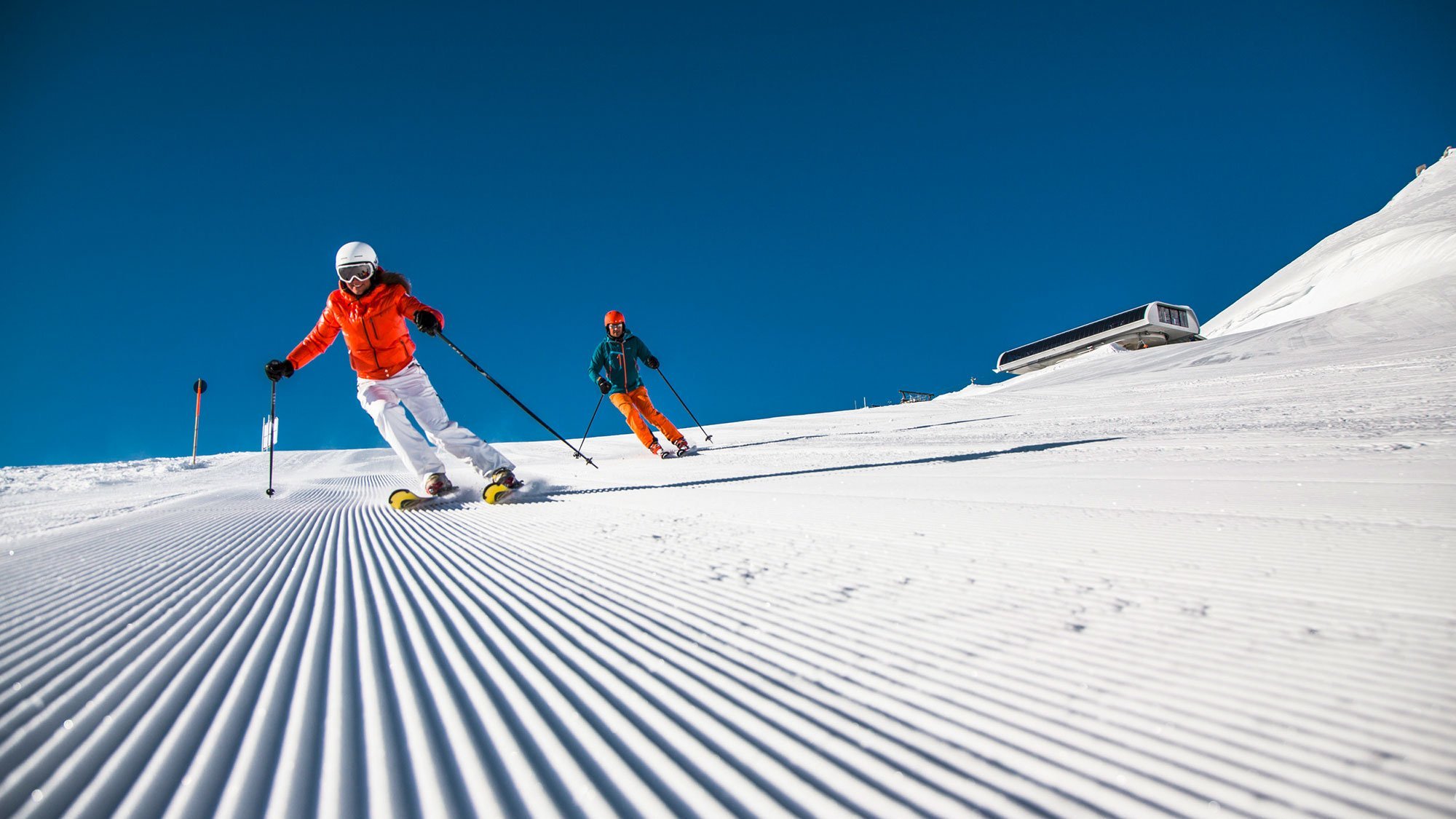 Zwei Skifahrer auf einer präparierten Piste unter klarem Himmel mit Bergen im Hintergrund.