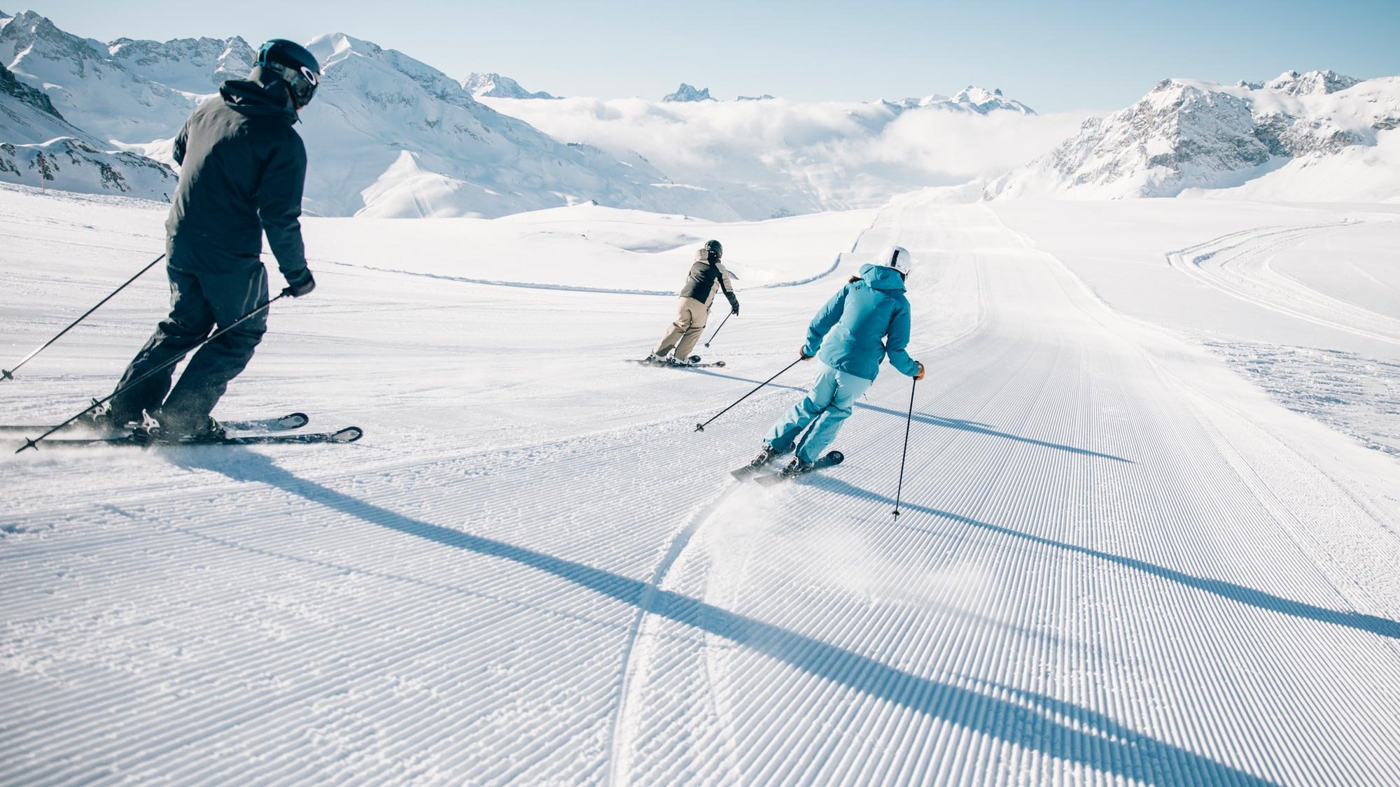Drei Skifahrer fahren eine schneebedeckte Piste in den Bergen hinunter unter einem klaren blauen Himmel.