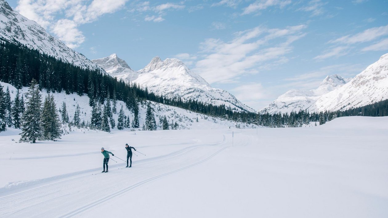 Zwei Personen beim Langlaufen in verschneiter Landschaft mit Bergen und Bäumen unter blauem Himmel